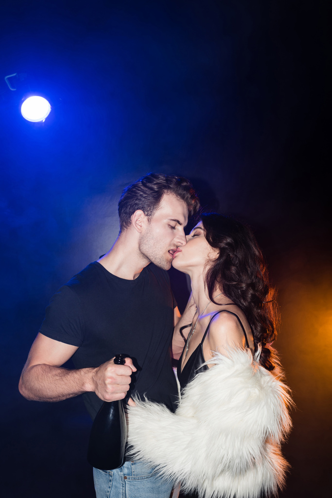 Mulher sedutora beijando namorado com garrafa de champanhe com retroiluminado em preto - Foto, Imagem