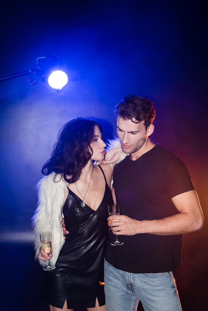 Lockige brünette Frau mit Glas Champagner lehnt an Freund im Hemd mit Hintergrundbeleuchtung auf schwarz - Foto, Bild