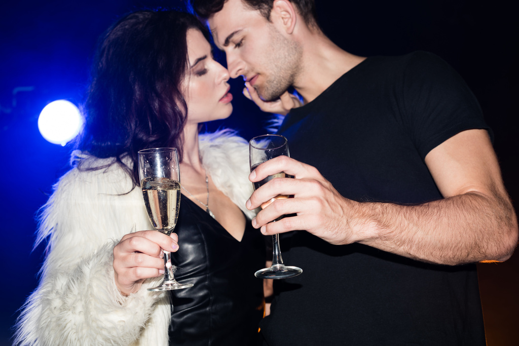 Verleidelijke vrouw met glas champagne aanraken vriendje kin met achtergrondverlichting op zwart - Foto, afbeelding