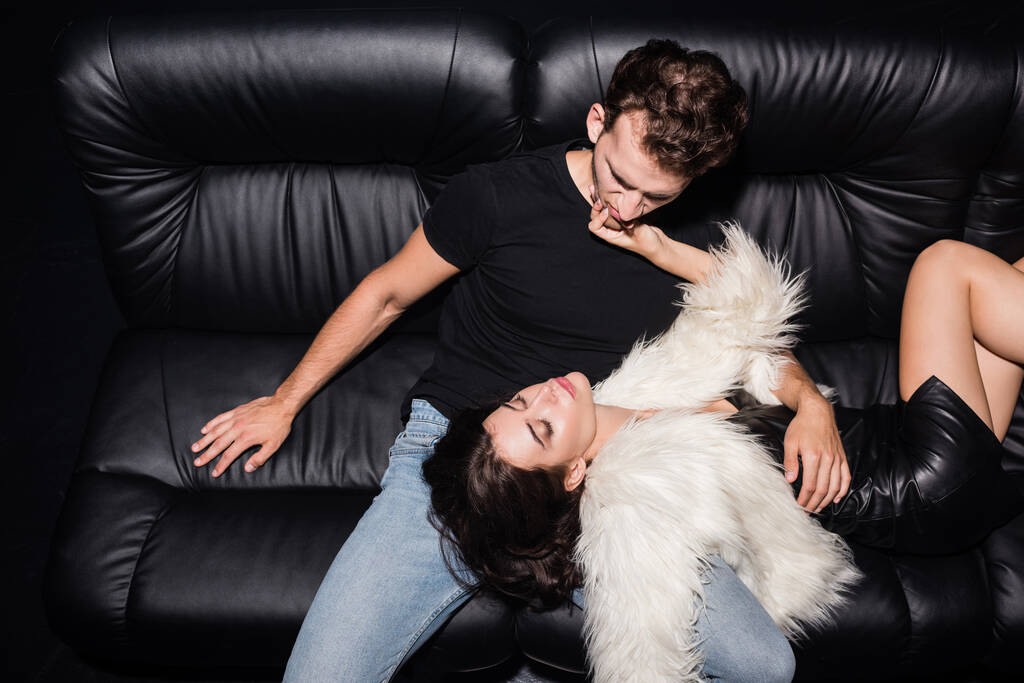 Vášnivá brunetka žena v bílém saku dotýkající přítel bradu, zatímco leží na pohovce v nočním klubu - Fotografie, Obrázek