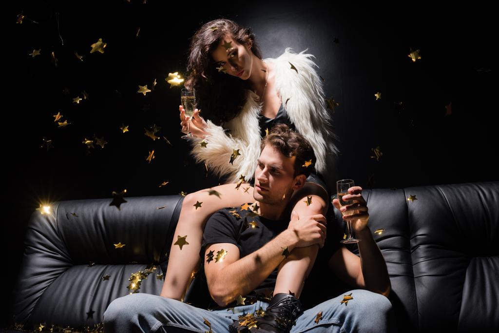 Сексуальная девушка смотрит в камеру с бокалом шампанского, сидя за мужчиной на диване с падающими конфетти в ночном клубе - Фото, изображение