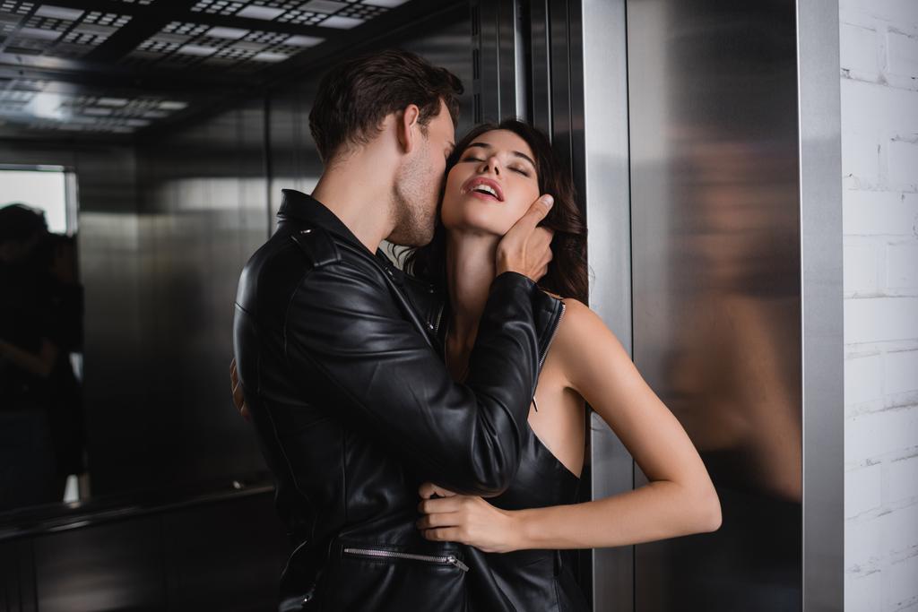 Мужчина в кожаной куртке касается шеи чувственной женщины с закрытыми глазами в лифте - Фото, изображение