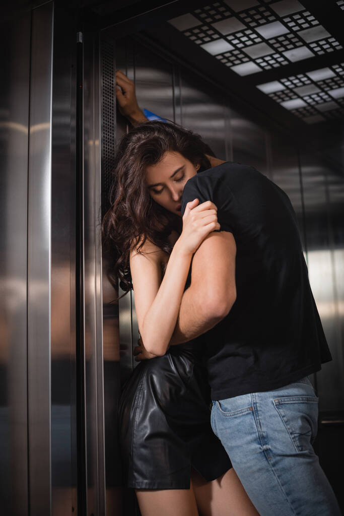 Szenvedélyes férfi fekete pólóban és farmerban, szexi nőt ölel át csukott szemmel a lift bejáratánál. - Fotó, kép