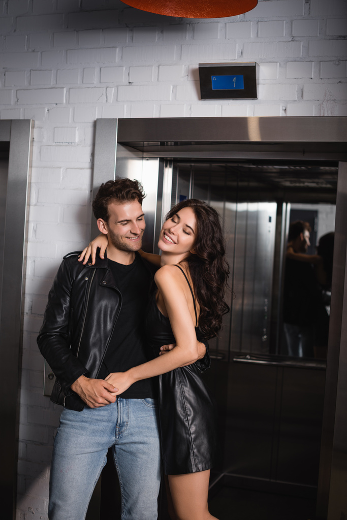 Fröhliche Frau mit lockigem Haar umarmt lächelnden Mann in Lederjacke in der Nähe des Fahrstuhleingangs - Foto, Bild