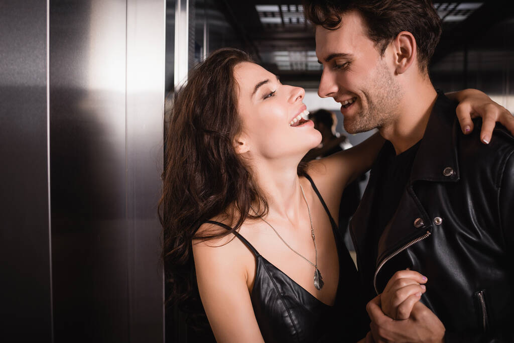 Μελαχρινή γυναίκα γελάει, ενώ αγκαλιάζει και κοιτάζοντας χαμογελαστός άνθρωπος με δερμάτινο μπουφάν στο ασανσέρ - Φωτογραφία, εικόνα