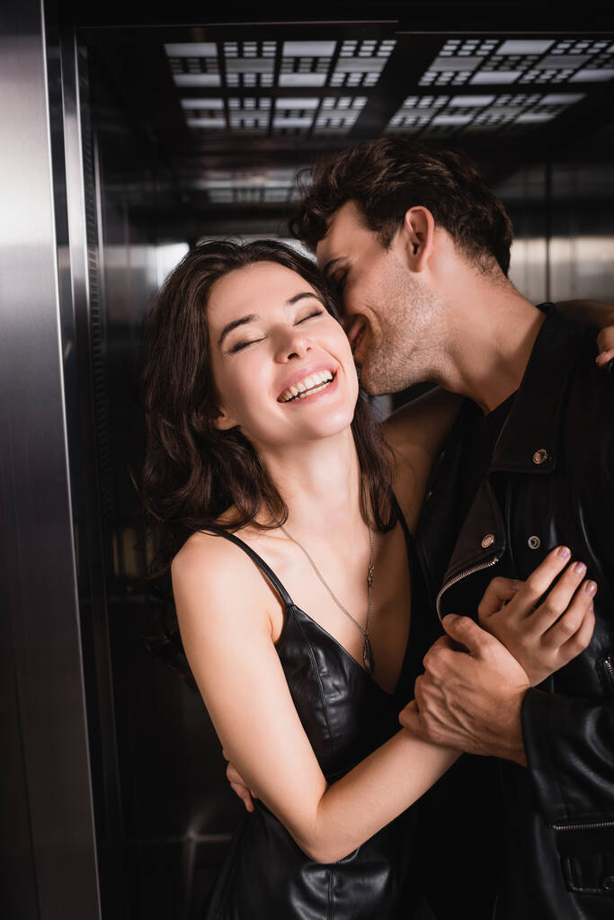 Hombre sonriente en chaqueta de cuero abrazando y cogido de la mano de mujer morena con los ojos cerrados riendo en ascensor - Foto, imagen