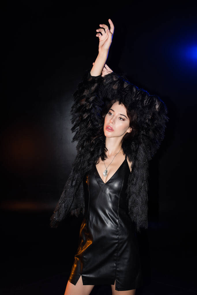 Szexi barna nő kezek a levegőben, visel bőr ruha és kabát, félrenéz, miközben áll a fekete - Fotó, kép