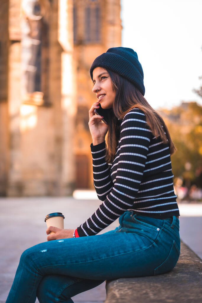 Életstílus, kaukázusi barna nő beszél telefonon a városban. Pihentető egy elvitelre kávé mellett egy gyönyörű templom a parkban. Fekete pulóver és gyapjúsapka - Fotó, kép