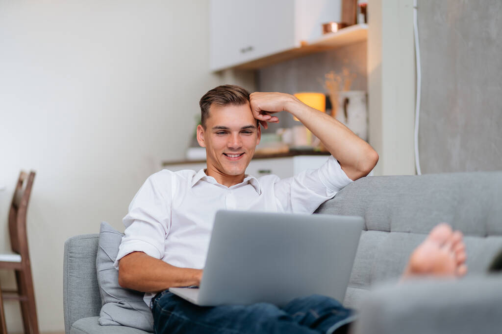 улыбчивый молодой человек просматривает Интернет, лежа на диване. - Фото, изображение