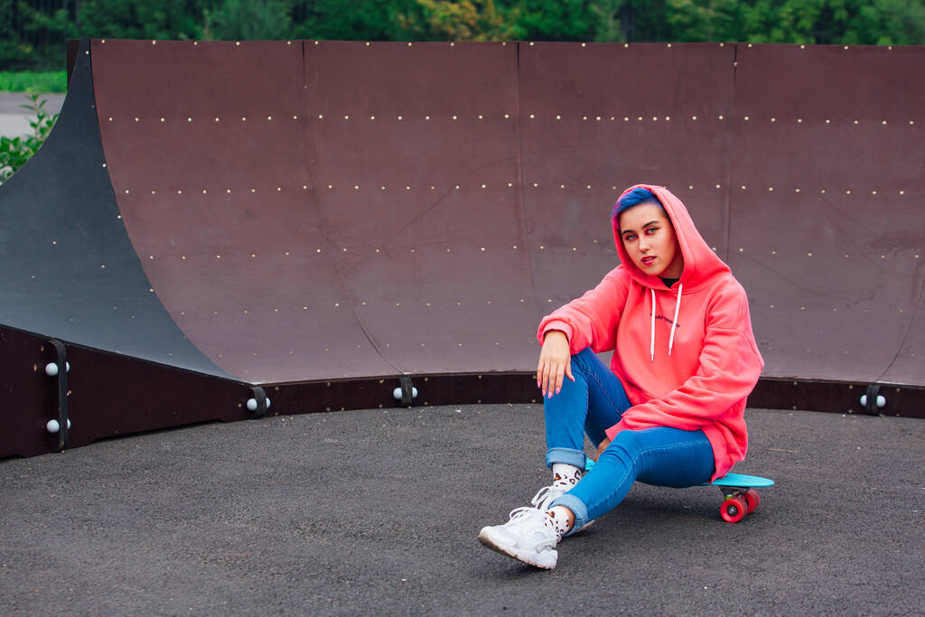 Stylish young woman sitting on her plastic skateboard in skatepark. Youth concept. - Photo, Image