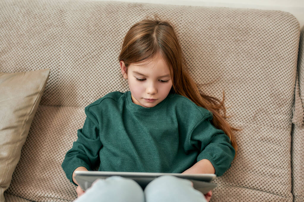 Une petite fille mignonne engagée dans un processus de jouer à des jeux sur une tablette tout en étant assis sur un canapé confortable - Photo, image