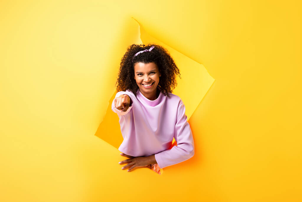 cheerful african american woman pointing with finger through ripped paper on yellow - Photo, Image