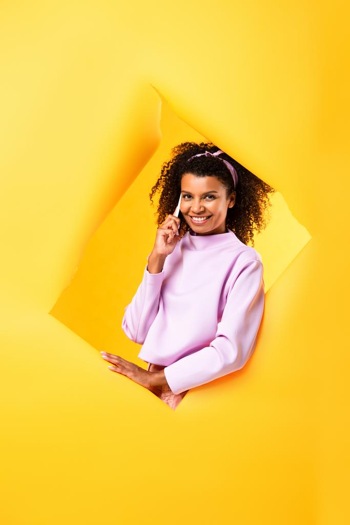 Весела афро-американська жінка розмовляє на смартфоні і дивиться на камеру через порвані папери на жовтому - Фото, зображення