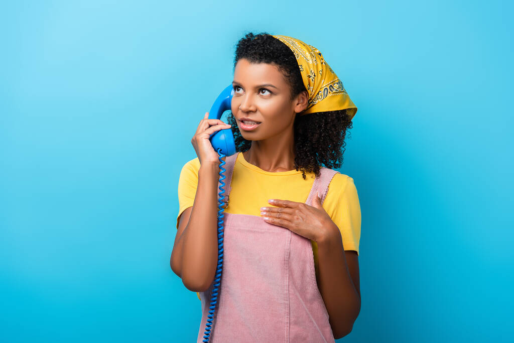 curly african american woman talking on retro telephone on blue - Photo, Image
