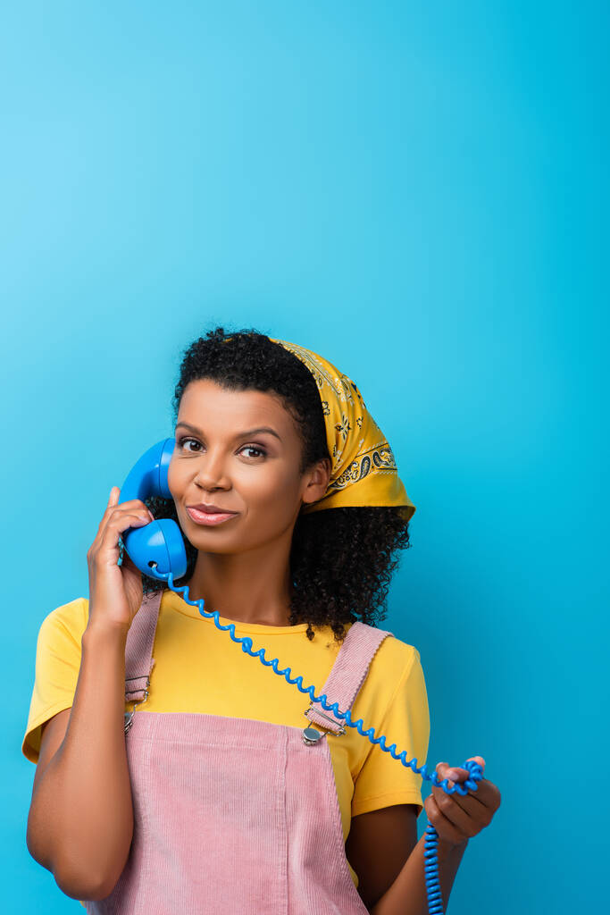 kudrnaté africké americká žena mluví na retro telefon a dívá se na kameru na modré - Fotografie, Obrázek