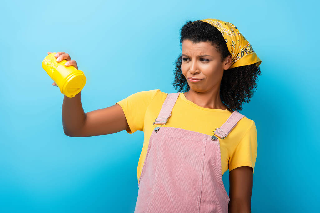 mujer afroamericana disgustada sosteniendo taza reutilizable vacía en azul - Foto, Imagen