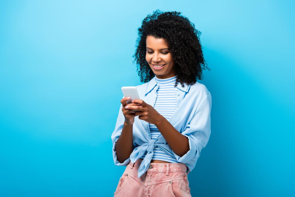 cheerful african american woman texting on smartphone on blue - Photo, Image