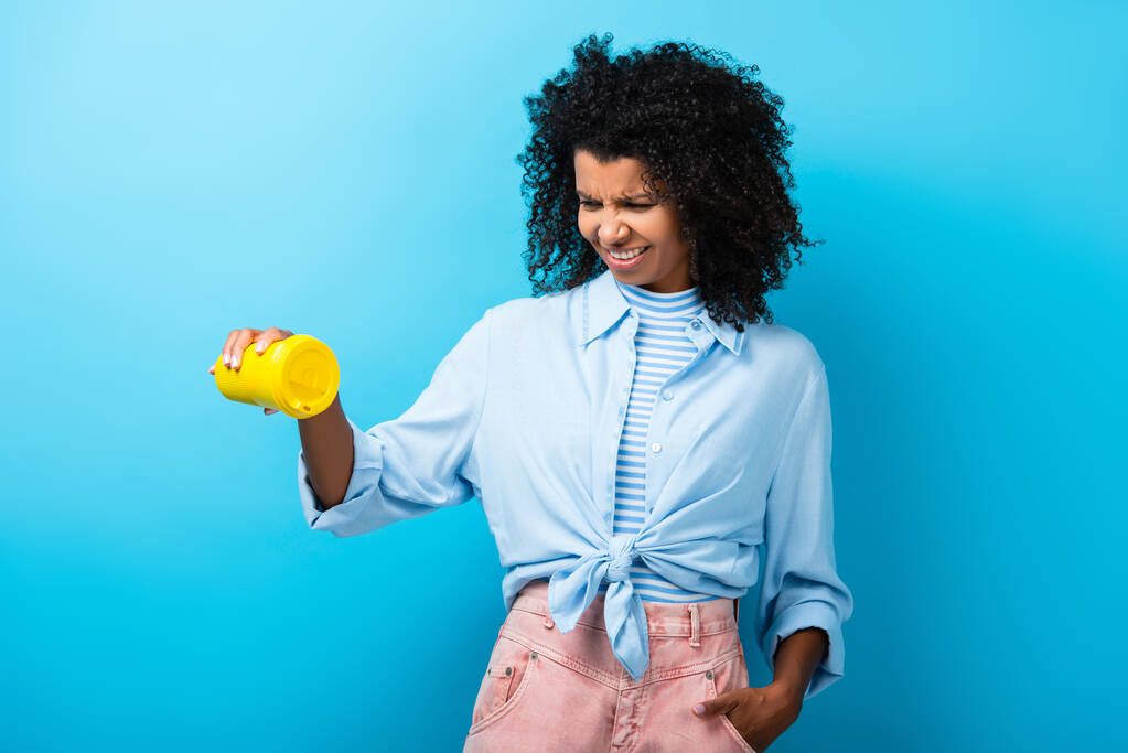 mujer afroamericana insatisfecha sosteniendo taza reutilizable vacía en azul - Foto, Imagen
