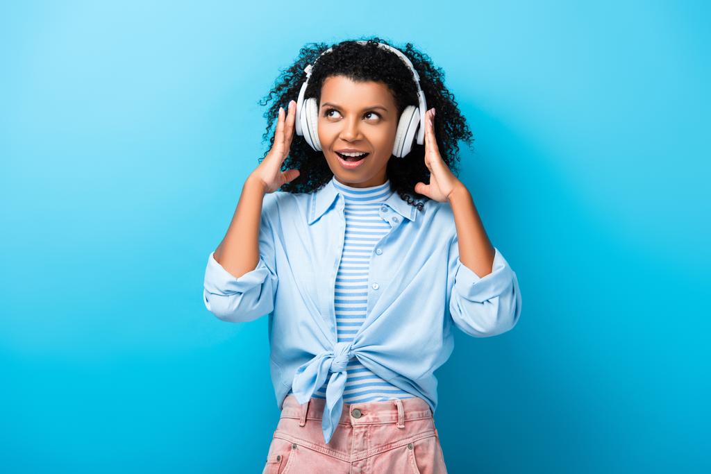 excited african american woman listening music in headphones on blue - Photo, Image