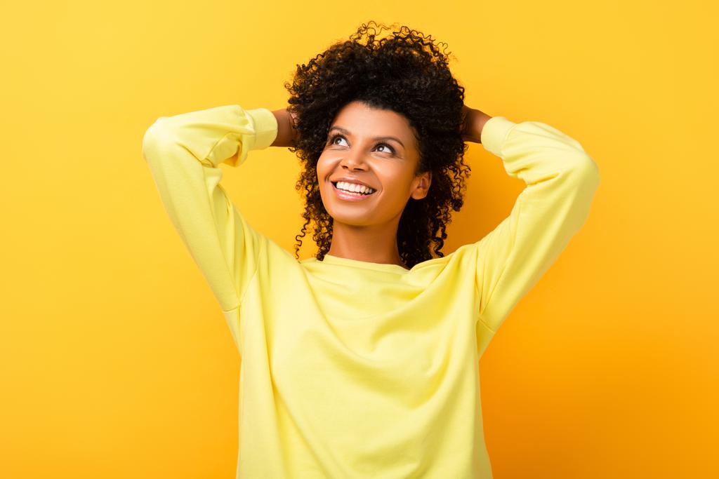 happy african american woman looking away while fixing curly hair on yellow - Photo, Image
