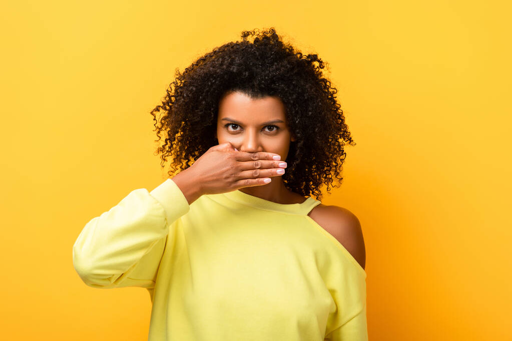 Αφροαμερικανή γυναίκα που καλύπτει το στόμα και κοιτάζει την κάμερα στο κίτρινο - Φωτογραφία, εικόνα
