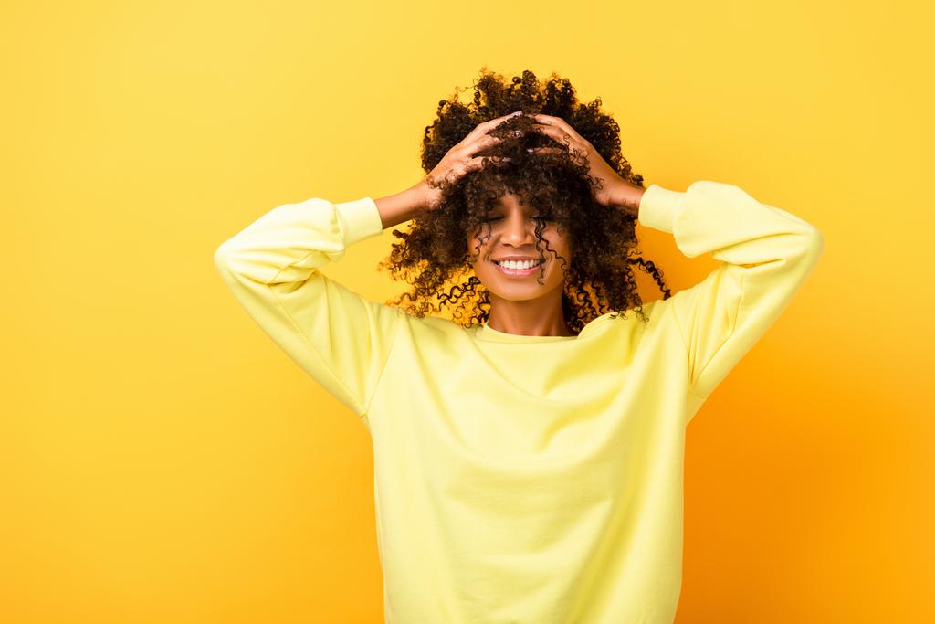 Χαρούμενη Αφροαμερικανή γυναίκα με κλειστά μάτια ενώ φτιάχνει σγουρά μαλλιά στο κίτρινο - Φωτογραφία, εικόνα