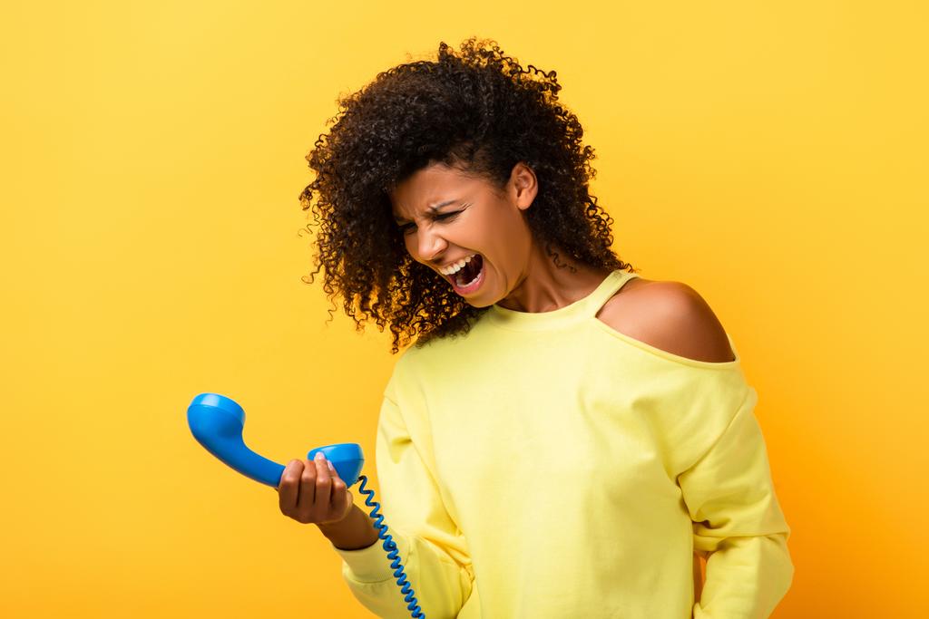 boze Afro-Amerikaanse vrouw op zoek naar vintage telefoon en schreeuwen op geel - Foto, afbeelding