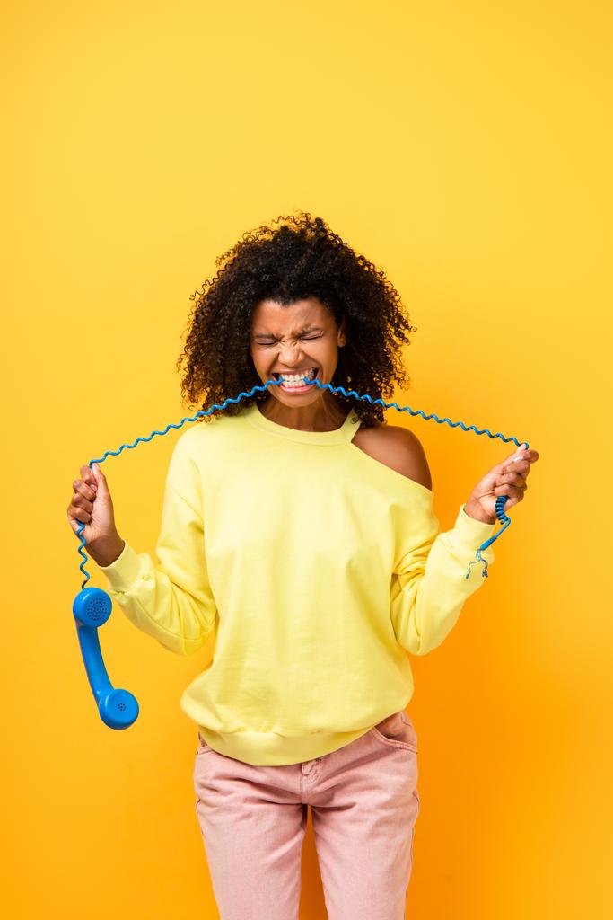 enojado africano americano mujer mordiendo cable de vintage teléfono en amarillo - Foto, Imagen