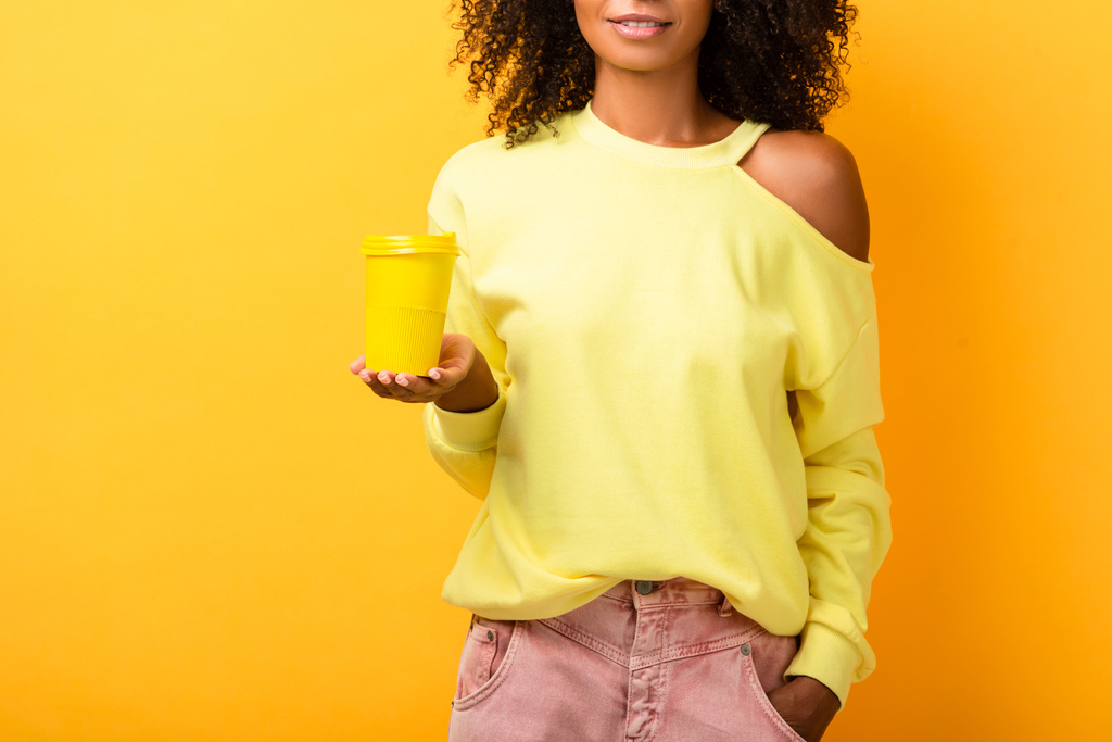 vista recortada de la mujer afroamericana sosteniendo taza reutilizable mientras está de pie con la mano en el bolsillo en amarillo - Foto, Imagen