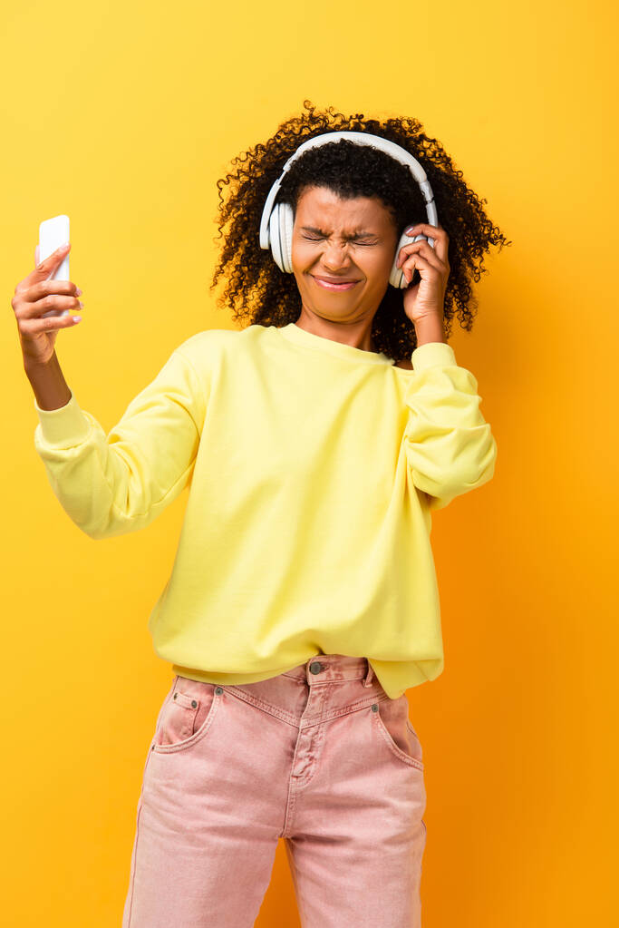 displeased african american woman in headphones holding smartphone on yellow - Photo, Image