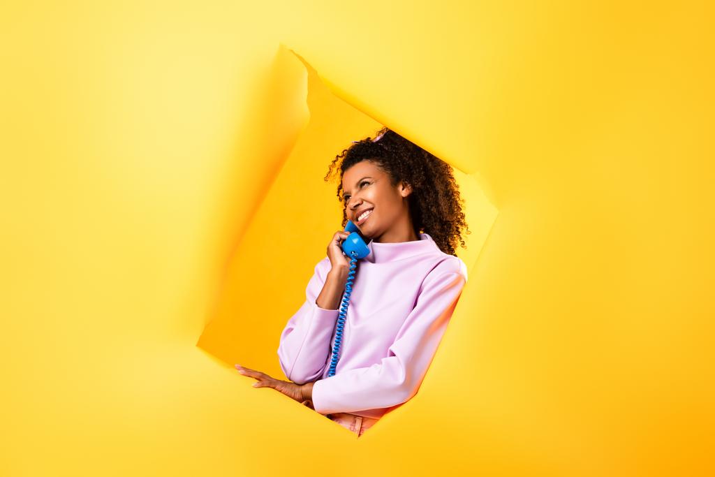 glücklich afrikanisch-amerikanische Frau spricht auf Retro-Telefon in der Nähe Loch in zerrissenem Papier auf gelbem Hintergrund  - Foto, Bild