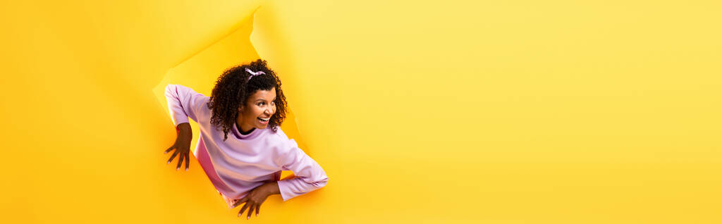 curiosa donna afroamericana che distoglie lo sguardo dal buco in carta strappata e sorride su sfondo giallo, striscione - Foto, immagini