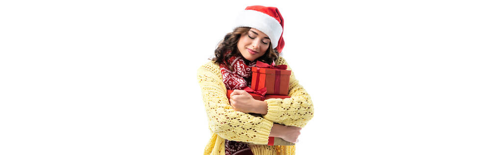 Panoramaaufnahme einer fröhlichen jungen Frau mit Weihnachtsmütze und Schal, die Geschenke isoliert auf Weiß hält - Foto, Bild