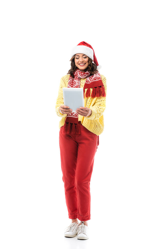 fröhliche junge Frau mit Weihnachtsmütze und Schal, die auf ein digitales Tablet schaut, das isoliert auf weißem Grund steht  - Foto, Bild