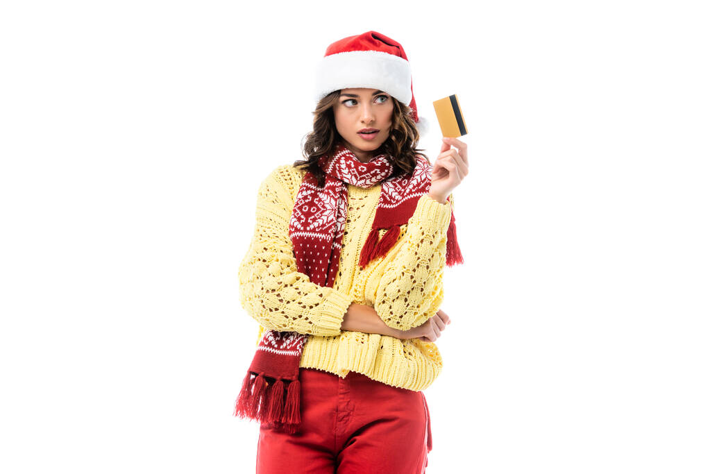 giovane donna in cappello di Babbo Natale e sciarpa guardando la carta di credito isolata su bianco  - Foto, immagini