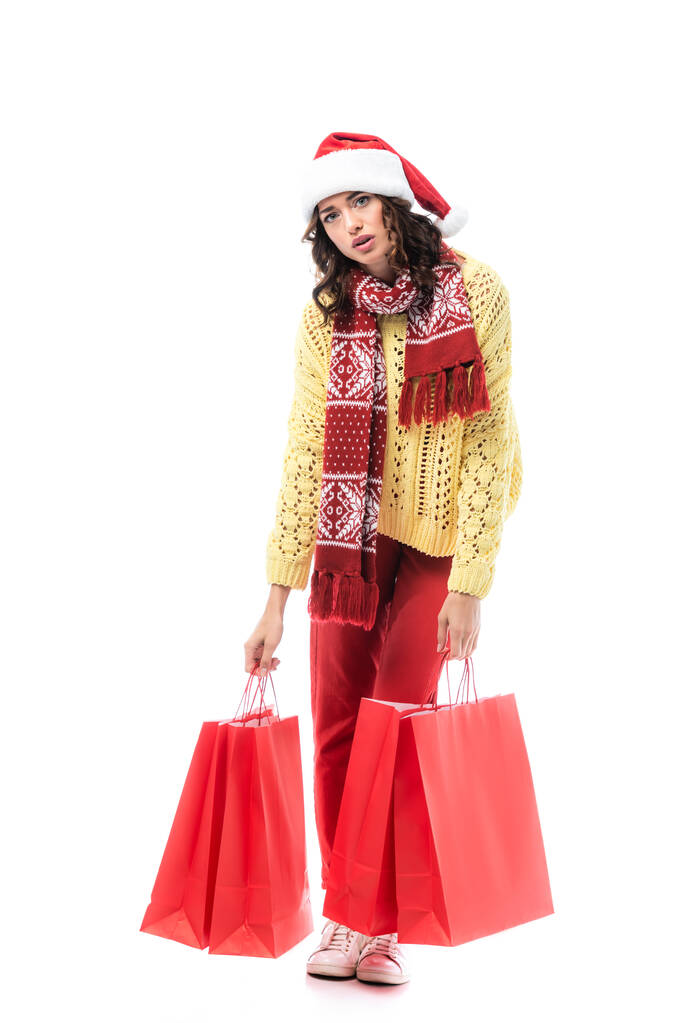 stanca giovane donna in cappello di Babbo Natale e sciarpa con ornamento in possesso di borse della spesa rosse isolate su bianco  - Foto, immagini