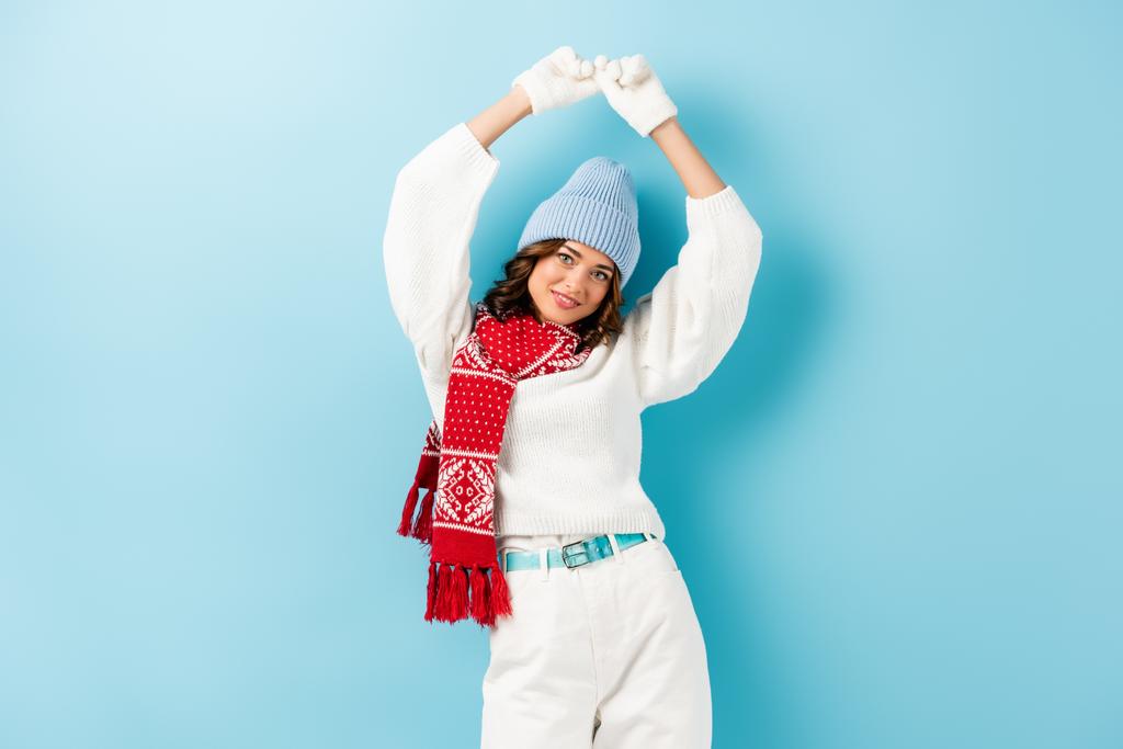 молодая женщина в зимнем наряде и аксессуары, стоящие с руками над головой на голубом - Фото, изображение