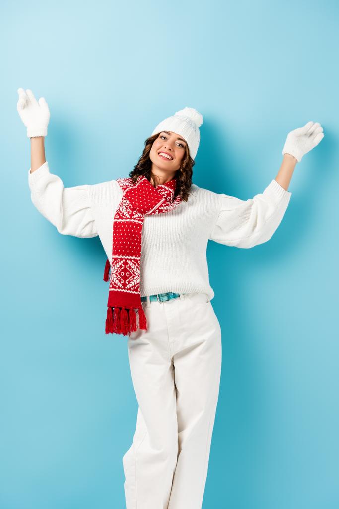 Радостная женщина в зимнем наряде, теплый шарф, перчатки и шляпа стоят с протянутыми руками на голубом - Фото, изображение