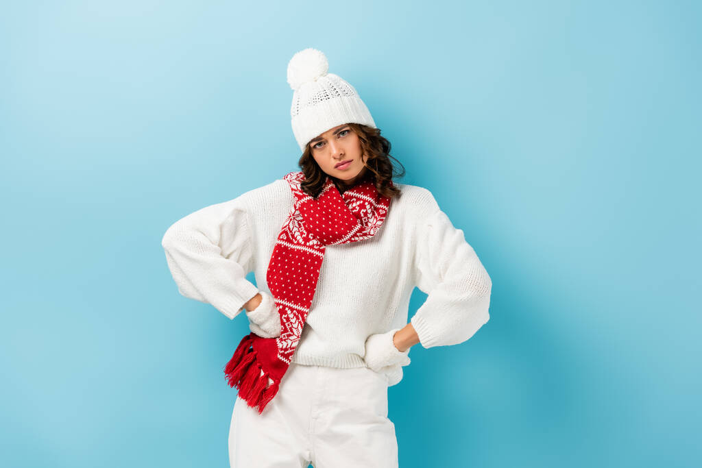 sérieuse jeune femme en tenue d'hiver blanche debout avec les mains sur les hanches sur bleu  - Photo, image