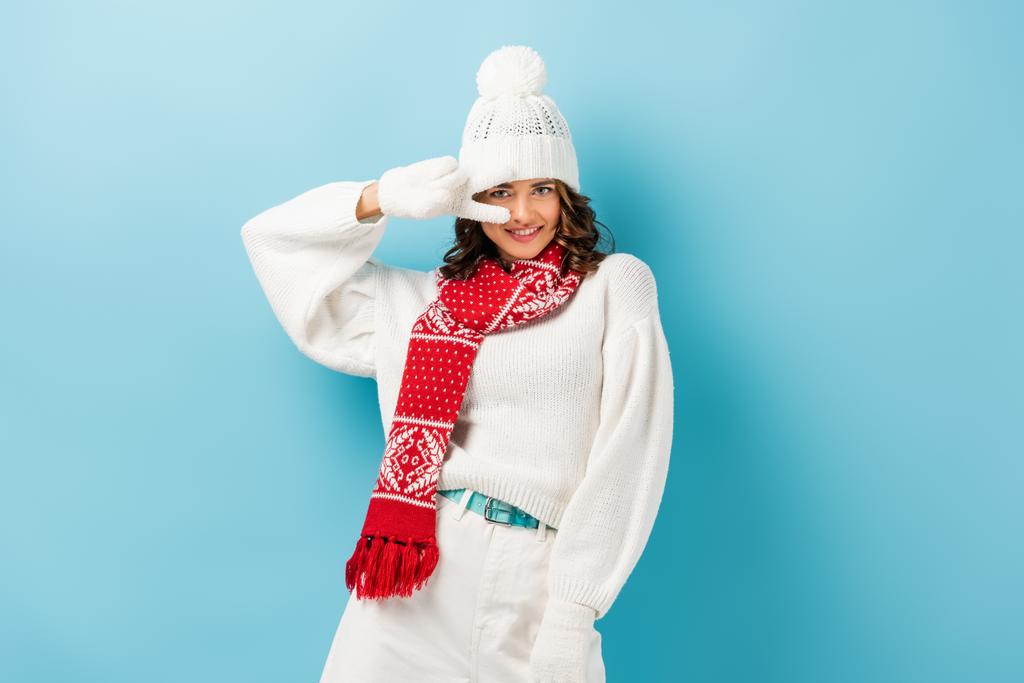 tevreden jonge vrouw in witte winter outfit tonen vrede teken op blauw  - Foto, afbeelding