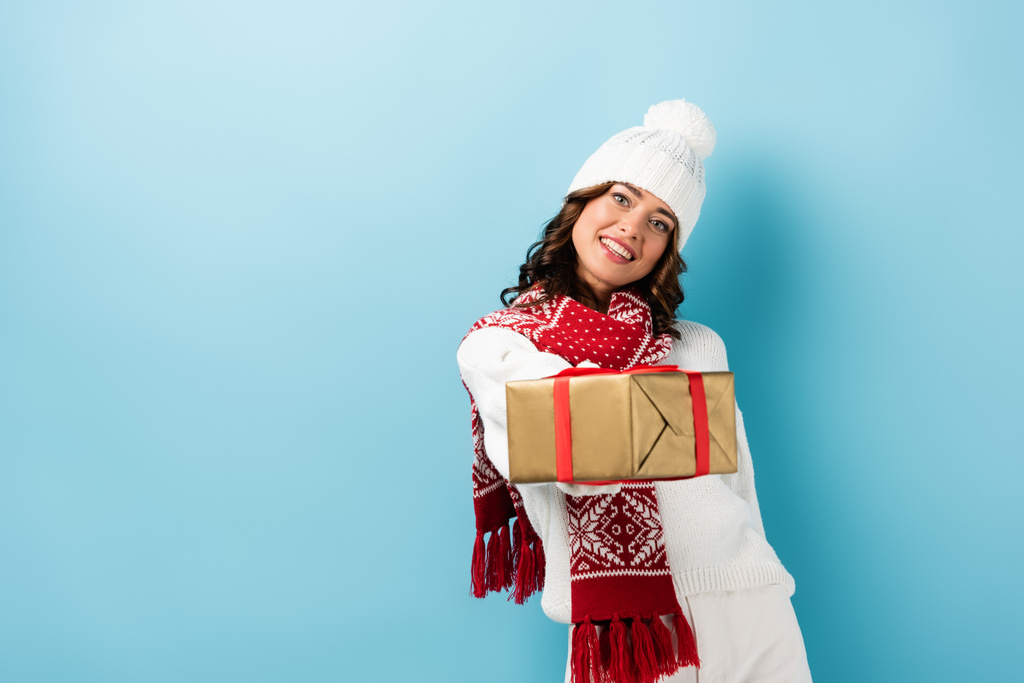 молодая довольная женщина в зимнем наряде держит завернутый подарок на голубом - Фото, изображение