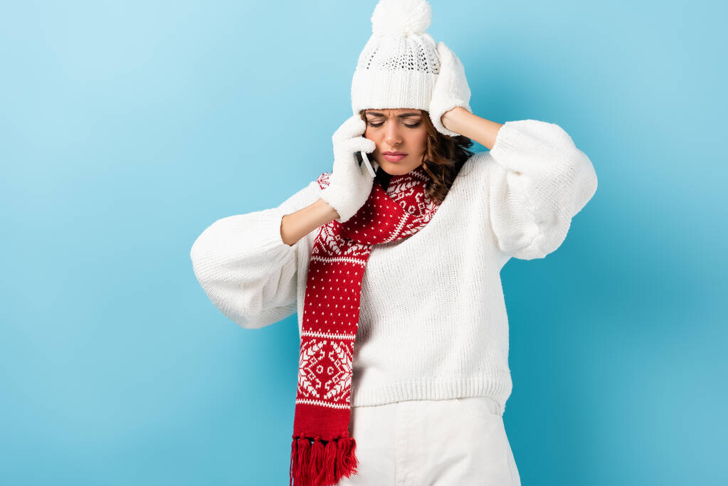 boos jonge vrouw in witte winter outfit praten op smartphone op blauw - Foto, afbeelding