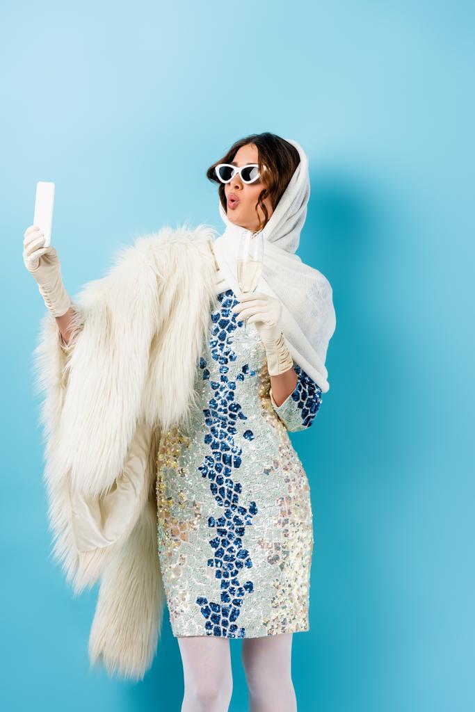 стильная женщина в солнечных очках держит бокал шампанского и делает селфи на голубом - Фото, изображение
