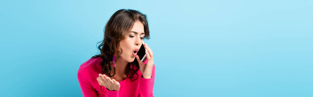 horizontaal gewas van brunette vrouw schreeuwen tijdens het praten op smartphone en gebaren op blauw - Foto, afbeelding