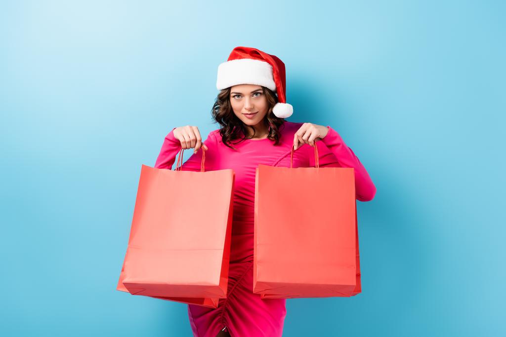 junge brünette Frau mit Weihnachtsmann-Hut hält Einkaufstüten in der Hand und blickt in die Kamera auf blau - Foto, Bild