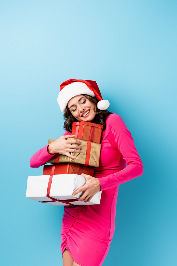młoda radosna kobieta w kapeluszu Mikołaja i sukience obejmującej zapakowane prezenty na niebiesko - Zdjęcie, obraz