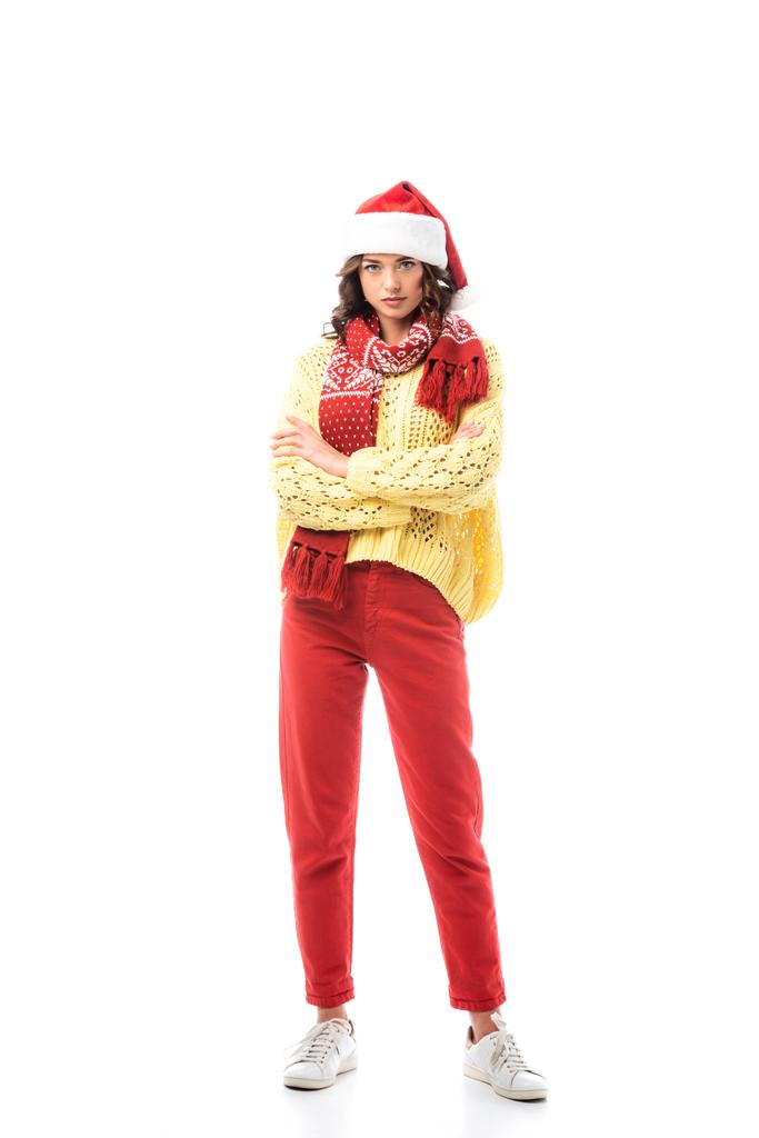 молодая брюнетка в шляпе Санты и красный шарф стоя со скрещенными руками изолированы на белом  - Фото, изображение