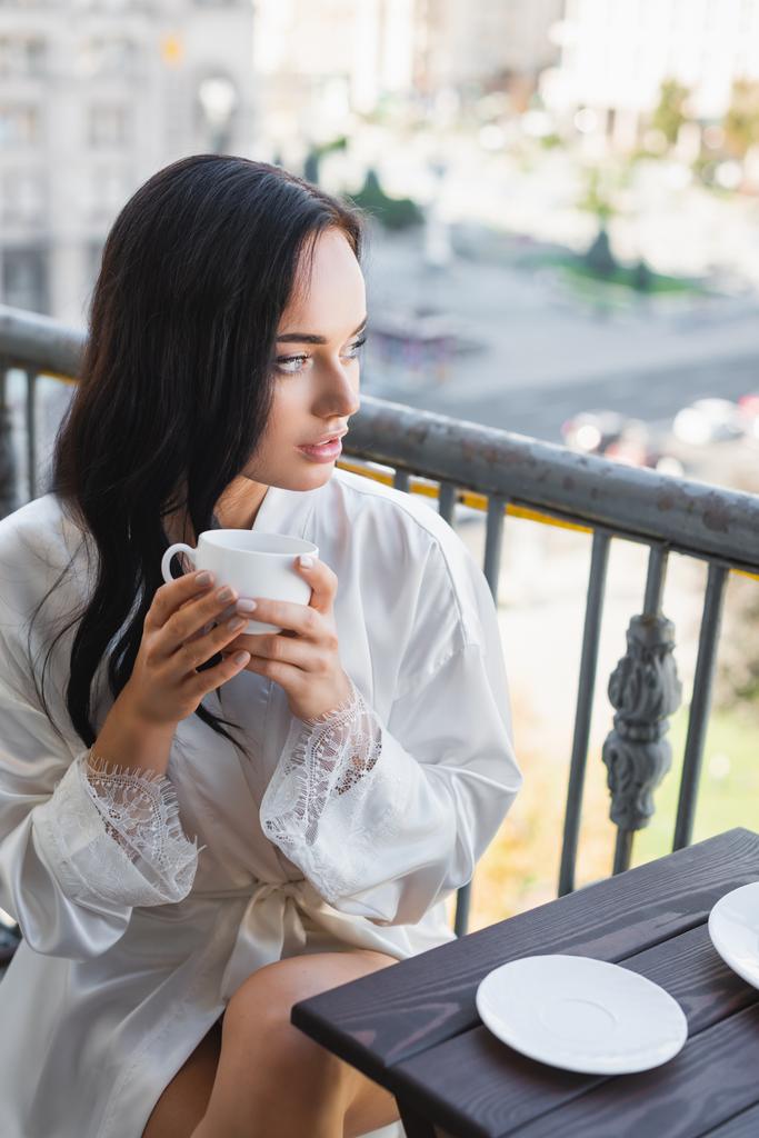 красивая брюнетка в белом халате пьет кофе на балконе - Фото, изображение