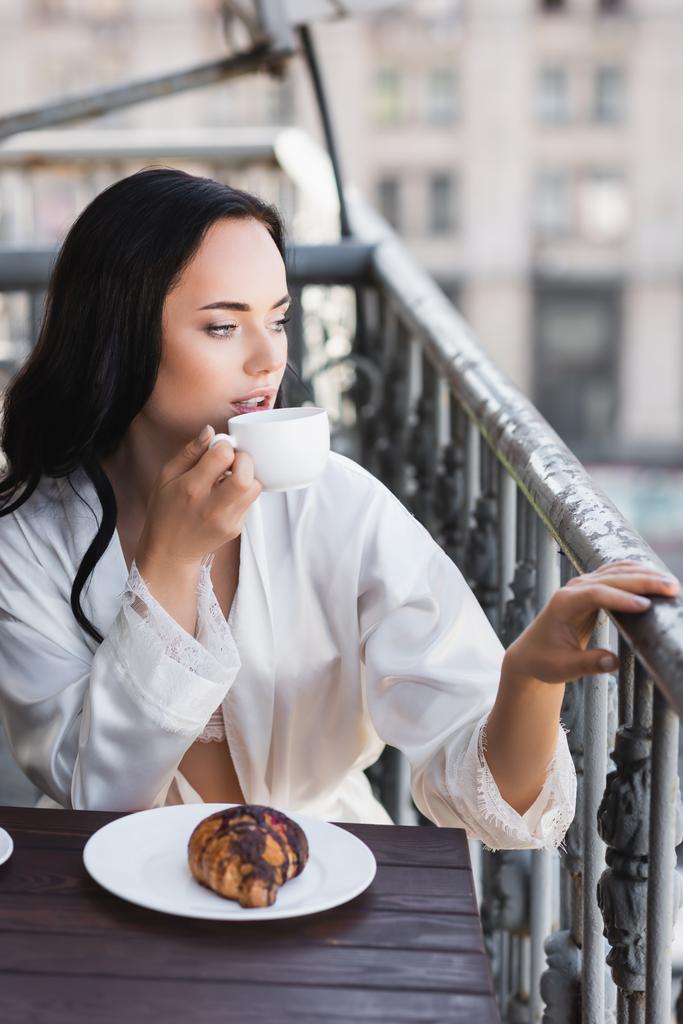 όμορφη μελαχρινή γυναίκα με λευκή ρόμπα πίνοντας καφέ και κοιτάζοντας μακριά - Φωτογραφία, εικόνα