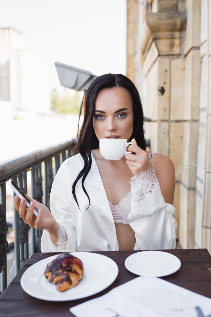 belle femme brune en robe blanche tenant smartphone et prenant le petit déjeuner sur le balcon - Photo, image